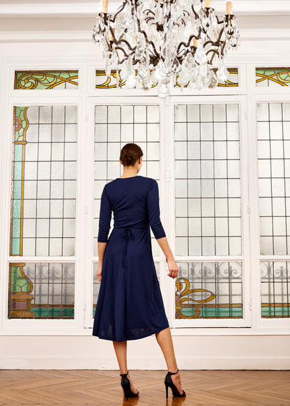 Woman models navy blue tea-length dress in front of windows